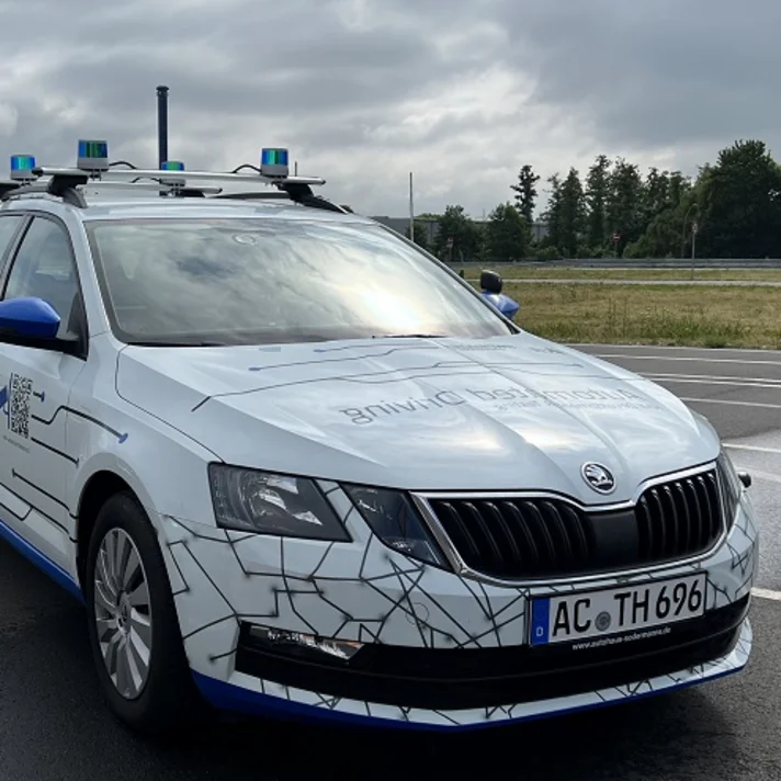 Autonomes Forschungsfahrzeug auf Straße mit Sensorik auf dem Dach. Es ist weiß mit schwarzer Linienmusterung.