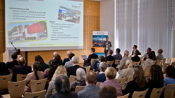 Vortragender steht vor Publikum in einem Konferenzraum mit Präsentationsfolie und Banner im Hintergrund