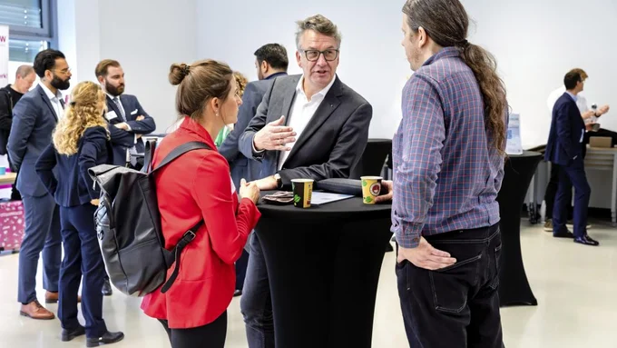 Mehrere Personen stehen in einem hellen Raum an Stehtischen, einige halten Kaffeebecher, eine Frau trägt einen schwarzen Rucksack und einen roten Blazer.