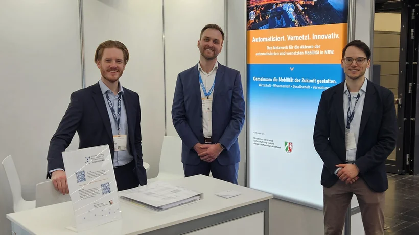 Drei Männer stehen am Messestand von innocam.NRW auf der XPONENTIAL 2025 und präsentieren Informationsmaterial zur vernetzten Mobilität.