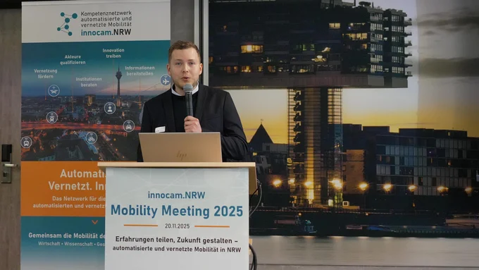 Person steht an einem Rednerpult mit Mikrofon und Laptop, im Hintergrund Banner und großes Bild eines modernen Gebäudes bei Dämmerung.