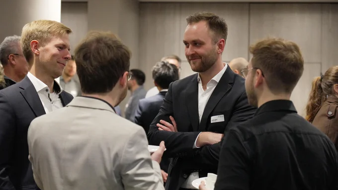 Mehrere Personen in Businesskleidung unterhalten sich in einem Innenraum bei einer Veranstaltung, im Vordergrund vier Männer.