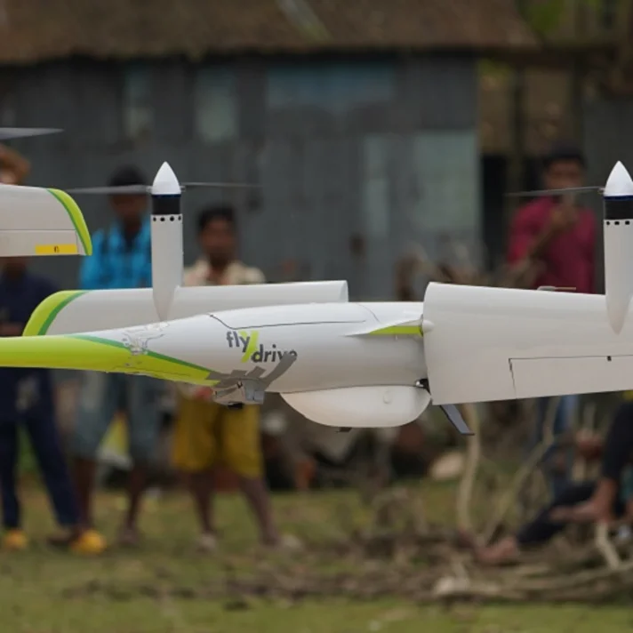 Eine grün-weiße Drohne fliegt bodennah in einem ländlichen Gebiet, im Hintergrund stehen mehrere Menschen.