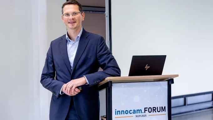 Mann in dunkelblauem Anzug lehnt an Rednerpult mit Laptop, auf dem Schild steht 'innocam.FORUM 18.09.2025'.