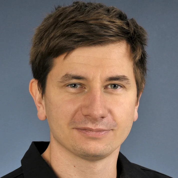 Portrait von Manuel Cymbaly zeigt in in einem dunklen Hemd, lächelnd mit dunklem Haar vor einem dunkelblauen Hintergrund.