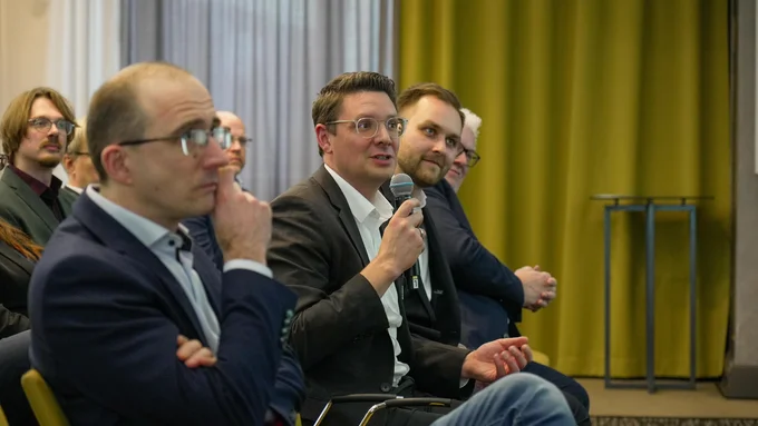 Mehrere Personen sitzen in den Zuschauerrängen eines Konferenzraumes, eine Person hält ein Mikrofon und spricht, im Hintergrund ein gelber Vorhang und ein kleiner Tisch.