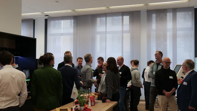 Gruppe von Menschen in einem hellen Raum mit großen Fenstern, die sich unterhalten und an einem Tisch mit Pflanzen und Getränken stehen