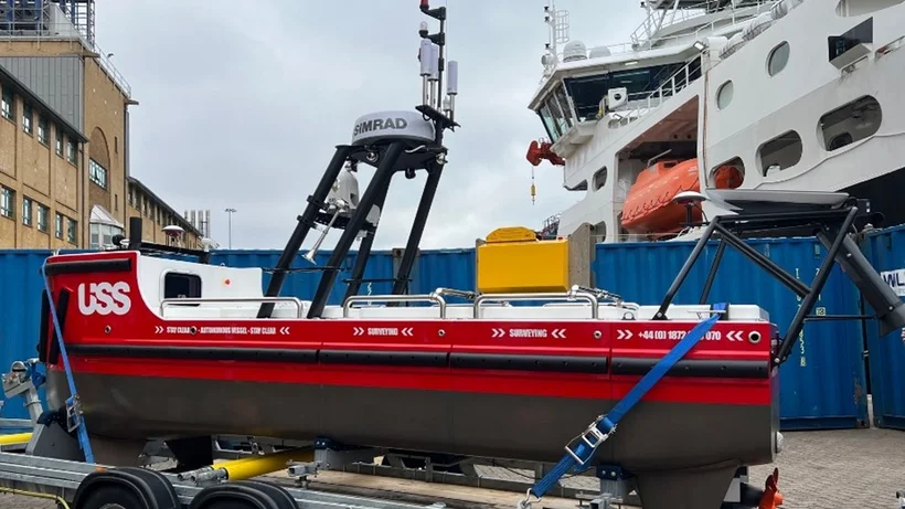 Ein kleines, rotes autonomes Schiff mit der Aufschrift 'USS' auf der Seite steht auf einem Anhänger. Das Schiff ist mit verschiedenen technischen Geräten ausgestattet, darunter eine Antenne mit der Aufschrift 'SIMRAD'. Im Hintergrund sind ein größeres weißes Schiff und ein Gebäude zu sehen. Das autonome Schiff ist mit blauen Gurten auf dem Anhänger gesichert.