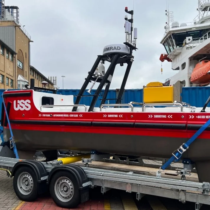Ein kleines, rotes autonomes Schiff mit der Aufschrift 'USS' auf der Seite steht auf einem Anhänger. Das Schiff ist mit verschiedenen technischen Geräten ausgestattet, darunter eine Antenne mit der Aufschrift 'SIMRAD'. Im Hintergrund sind ein größeres weißes Schiff und ein Gebäude zu sehen. Das autonome Schiff ist mit blauen Gurten auf dem Anhänger gesichert.