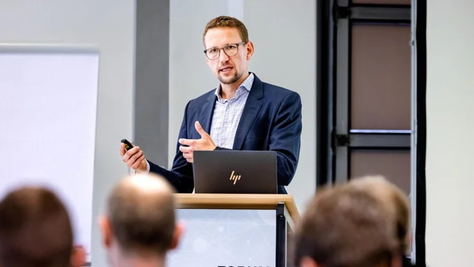 Mann in dunklem Sakko und hellblauem Hemd hält Präsentation am Rednerpult mit Laptop, Publikum im Vordergrund, Veranstaltung innocam.FORUM.