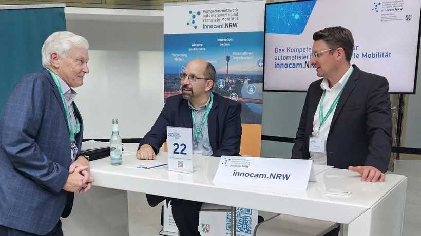 Drei Männer in Anzugjacken stehen und sitzen an einem Messestand des Netzwerks ‘innocam.NRW’. Sie unterhalten sich freundlich an einem hohen weißen Tisch, auf dem Informationsmaterial, eine Flasche Wasser und ein Tischaufsteller mit der Nummer 22 stehen. Hinter ihnen befinden sich Roll-up-Banner und ein Monitor mit Informationen über automatisierte und vernetzte Mobilität.