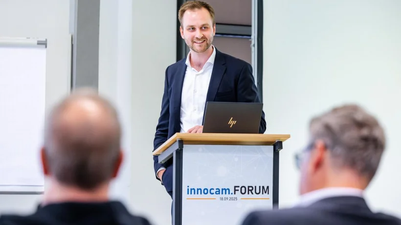 Ein Mann in dunklem Anzug und weißem Hemd steht hinter einem Rednerpult mit Laptop und dem Schriftzug 'innocam.FORUM 18.09.2025'. Im Vordergrund sind zwei Männer von hinten zu sehen.