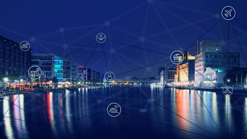 Nächtliche, beleuchtete Panoramaansicht der neunen Innenhafens der Stadt Duisburg im dunklen Blauton mit Lichtakzenten, mit modernen Gebäuden an den Ufern rechts und links, deren Licht sich im Wasser spiegelt. Im Vordergrund sind Icons der automatisierten und vernetzten Mobilität zu sehen, die mit Linien Netzwerk miteinander verbunden sind. 