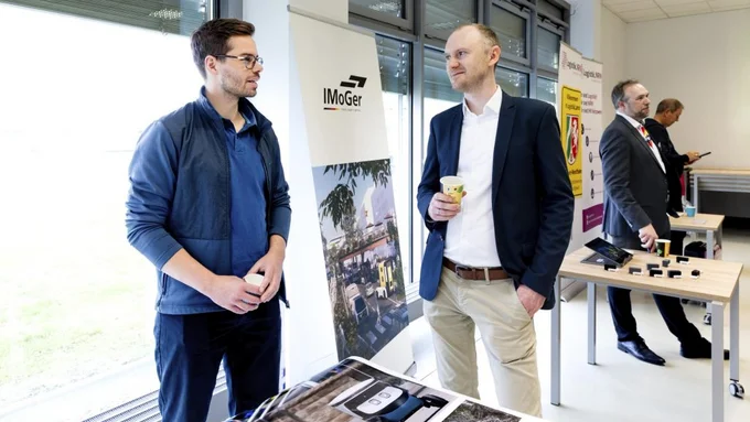 Zwei Männer stehen an einem Messestand mit Bannern der Marke 'ImoGer' und Informationsmaterial auf einem Tisch, im Hintergrund weitere Personen an einem zweiten Stand.