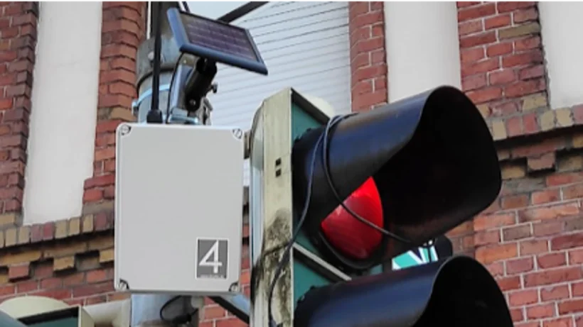 Rote Ampel mit montierter Sensorbox und Solarpanel an einem Gebäude.