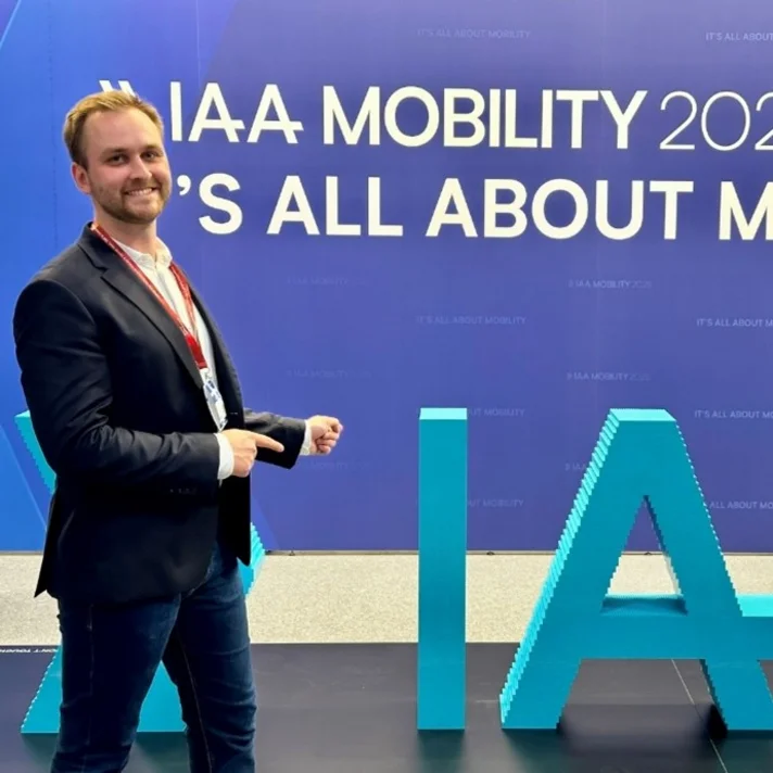 Ein junger, lächelnder Mann im Anzug zeigt auf den als Skulptur aufgestellten Schriftzug IAA in türkis.