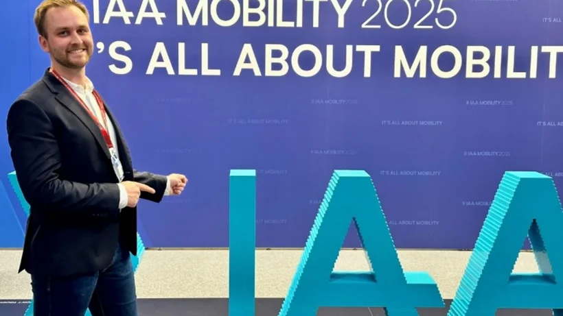 Ein junger, lächelnder Mann im Anzug zeigt auf den als Skulptur aufgestellten Schriftzug IAA in türkis.