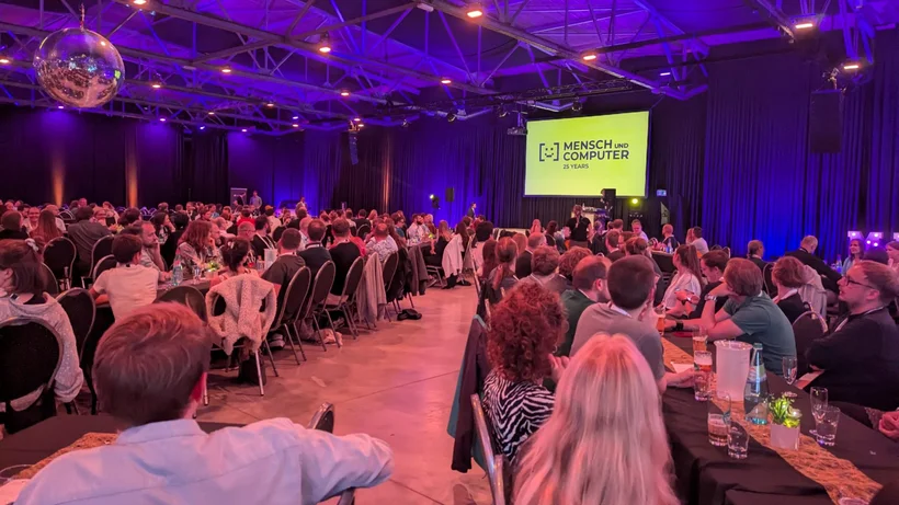 Eine Große Halle mit langen Tischreihen, an denen Personen mit Abendkleidung sitzen, der Raum ist bläulich beleuchtet und es ist eine Diskokugel aufgehangen, die PErsonen blicken auf eine Leinwand auf der Mensch und Computer Konferenz 2025 steht. 