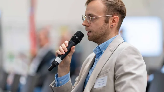 Ein junger Mann im hellen Sakko spricht in ein Mikrofon während einer Veranstaltung.