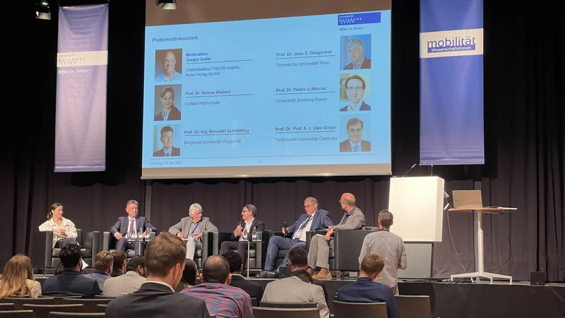 Foto zeigt aus Sicht des Publikums auf die Bühne. Auf der Bühne sitzen 7 Personen und eine Moderatorin für eine Paneldiskussion.