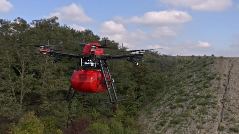 Rote Drohne mit sechs Rotoren fliegt über bewaldetes und bewachsenes Gelände
