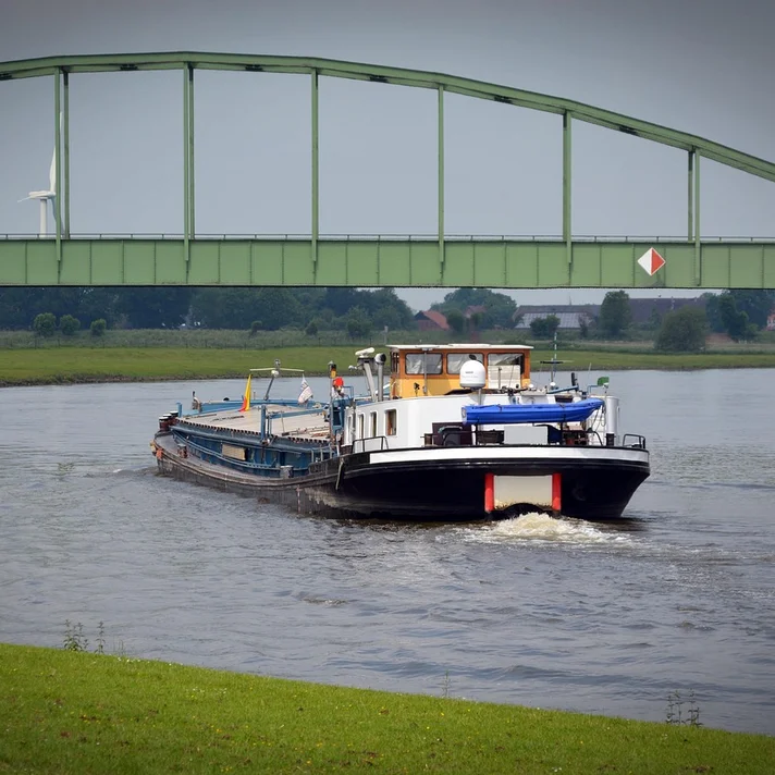 Binnenschiff mit weißem Aufbau und blauem Sonnenschutz fährt unter einer grünen Stahlbrücke auf einem Fluss.