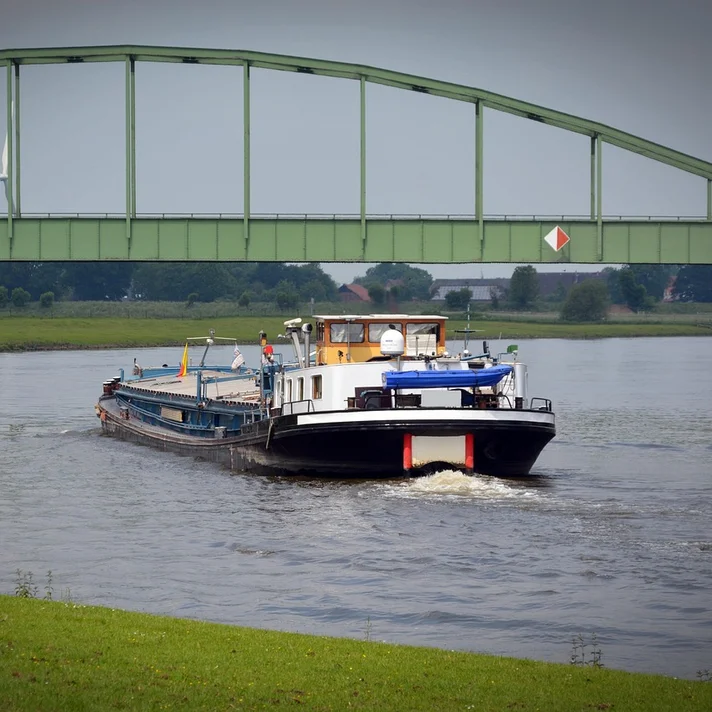 Binnenschiff mit weißem Aufbau und blauem Sonnenschutz fährt unter einer grünen Stahlbrücke auf einem Fluss.