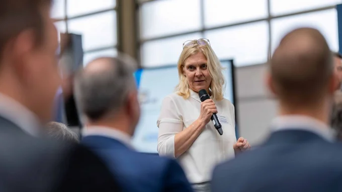 Frau in weißem Oberteil spricht in ein Mikrofon in einer Veranstaltungsszene.