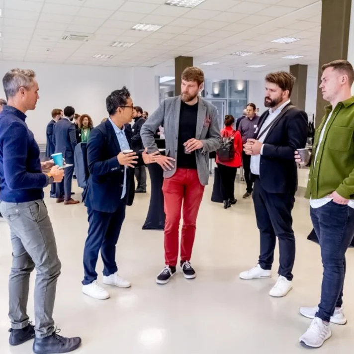 Mehrere Personen stehen in einem hellen, modernen Raum und unterhalten sich in kleinen Gruppen, einige halten Becher in der Hand.