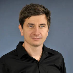 Portrait von Manuel Cymbaly zeigt in in einem dunklen Hemd, lächelnd mit dunklem Haar vor einem dunkelblauen Hintergrund.