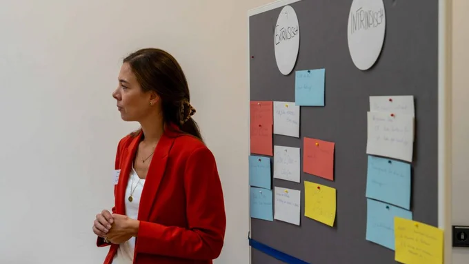 Frau im roten Blazer steht neben Pinnwand mit bunten Karten zu „extrinsischer“ und „intrinsischer“ Motivation.