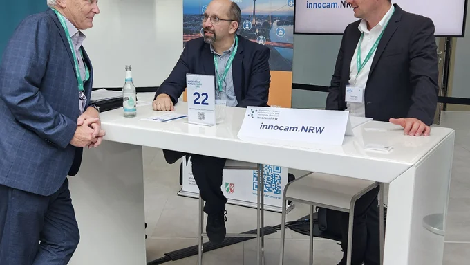 Drei Männer sprechen an einem Messestand mit dem Logo von innocam.NRW beim Innovationskongress 2025.