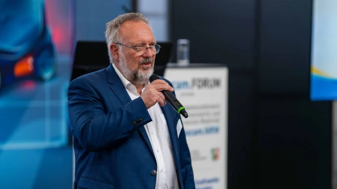 Ein älterer Mann im blauen Anzug spricht mit Mikrofon vor einem Messestand mit dem Logo „innocam.FORUM“.