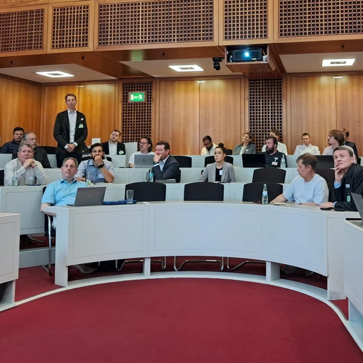 Blick in die Sitzreihen eines Hörsaals, roter Fußboden, graue besetzte ansteigende Reihen in einem engen Halbkreis, die PErsonen in den Reihen blicken aufmerksam auf eine Präsentaton. 