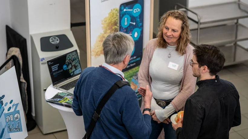 Drei Personen stehen an einem Stehtisch und sprechen über ein Projekt zur digitalen Stadtentwicklung mit einem Laptop und Infobroschüren.