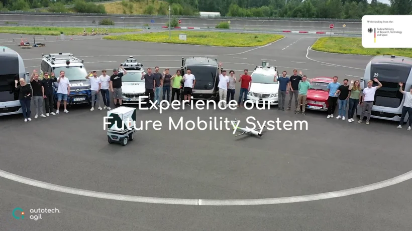 Eine Gruppe von etwa zwanzig Personen steht auf einem Testgelände neben verschiedenen Fahrzeugen – darunter autonome Busse, Vans, ein kleines Lieferfahrzeug und eine Drohne. In der Mitte steht der Text „Experience our Future Mobility System“. Unten links ist das Logo von „autotech.agil“ zu sehen, oben rechts das Logo des Bundesministeriums für Forschung und Technologie.