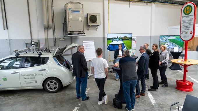 Gruppe steht an einem Infostand vor einem automatisierten Forschungsfahrzeug mit geöffneter Heckklappe.