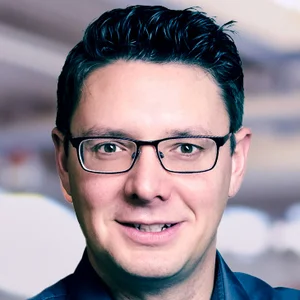 Porträt von Lasse Pipoh, Leiter der Geschäftsstelle, mit dunklem, kurz geschnittenem Haar, Brille und dunklem Hemd, vor unscharfem Hintergrund.