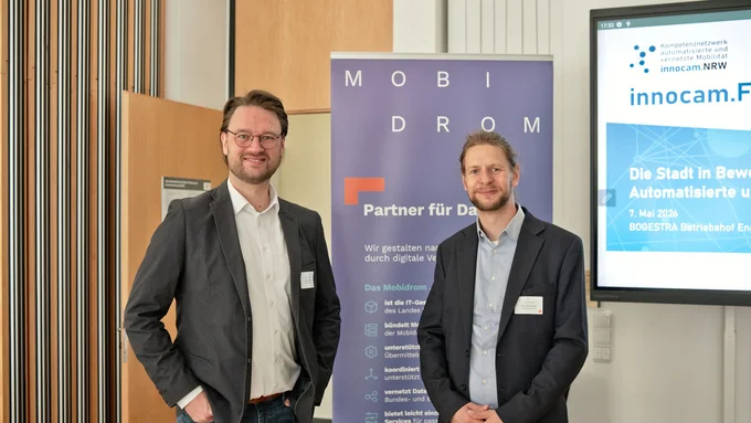 Zwei Männer in Anzügen stehen vor einem blauen Banner mit der Aufschrift 'MOBIDROM' und einem Bildschirm mit dem Logo 'innocam.NRW' und Text zur automatisierten Mobilität