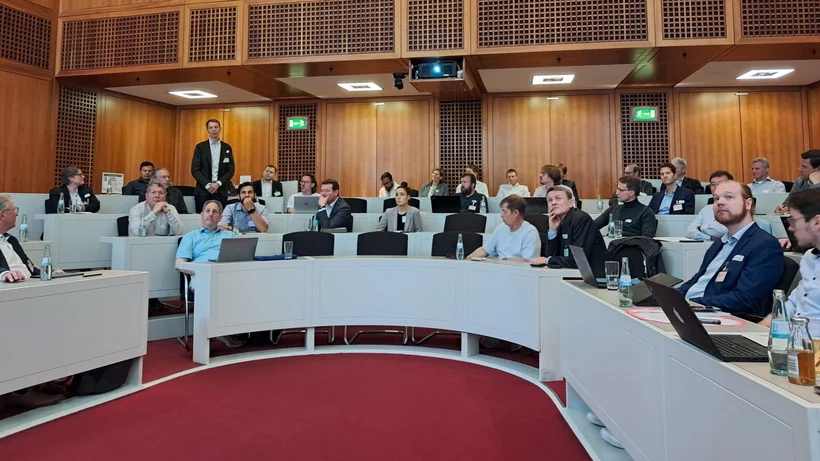 Mehrere Personen sitzen in einem Konferenzsaal und folgen einer Präsentation, während eine Person steht und spricht.