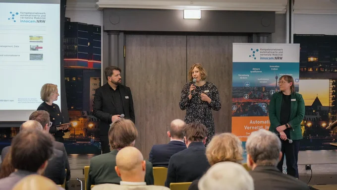 Vier Personen stehen vor einem Publikum in einem Konferenzraum, zwei Banner mit dem Logo und Text von innocam.NRW sind im Hintergrund sichtbar.