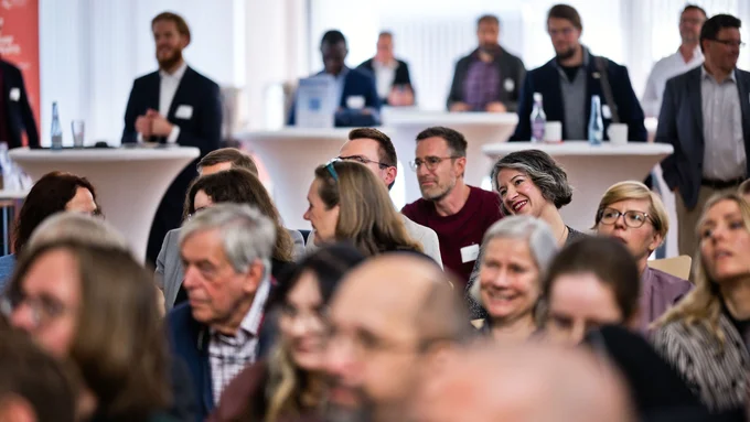 Gruppe von Menschen sitzt und steht in einem hellen Raum bei einer Veranstaltung