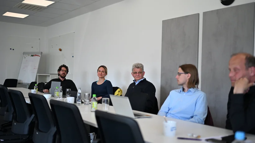 Fünf Personen sitzen an einem langen Konferenztisch in einem Besprechungsraum mit weißen Wänden und grauen Wandpaneelen
