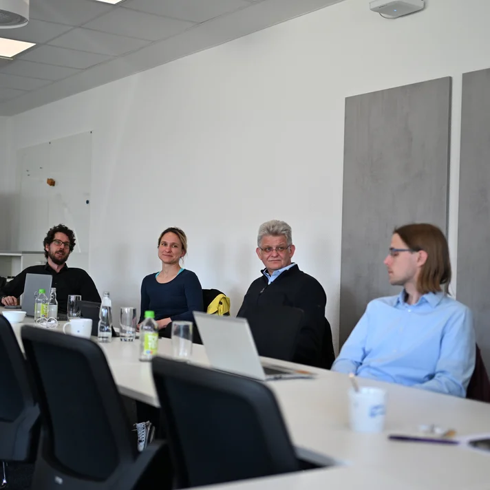 Fünf Personen sitzen an einem langen Konferenztisch in einem Besprechungsraum mit weißen Wänden und grauen Wandpaneelen