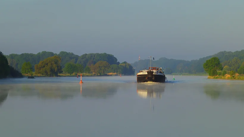 Ein Frachtschiff fährt in ruhigem, leicht nebligem Morgenlicht über einen breiten Fluss, umgeben von üppigem Grün und Bojen zur Fahrmarkierung.