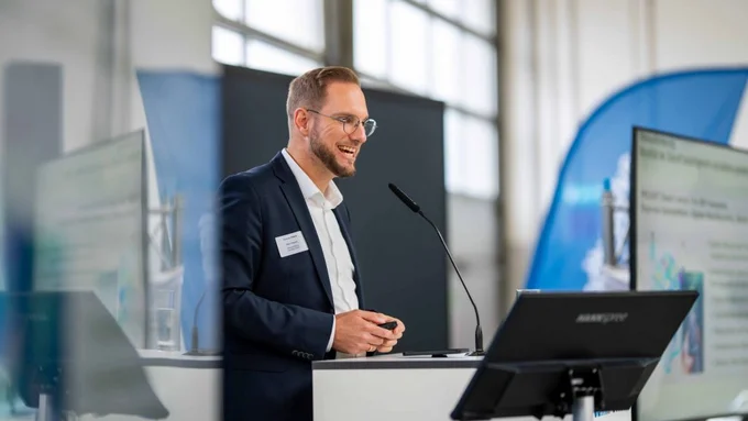 Mann mit Brille spricht lächelnd am Rednerpult mit Bildschirm und Präsentation im Hintergrund.