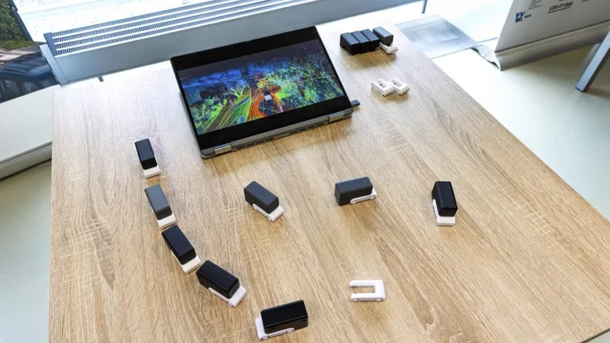 Auf einem Holztisch steht ein aufgeklappter Laptop mit bunter Landschaft auf dem Bildschirm, umgeben von mehreren kleinen schwarzen und weißen rechteckigen Objekten.