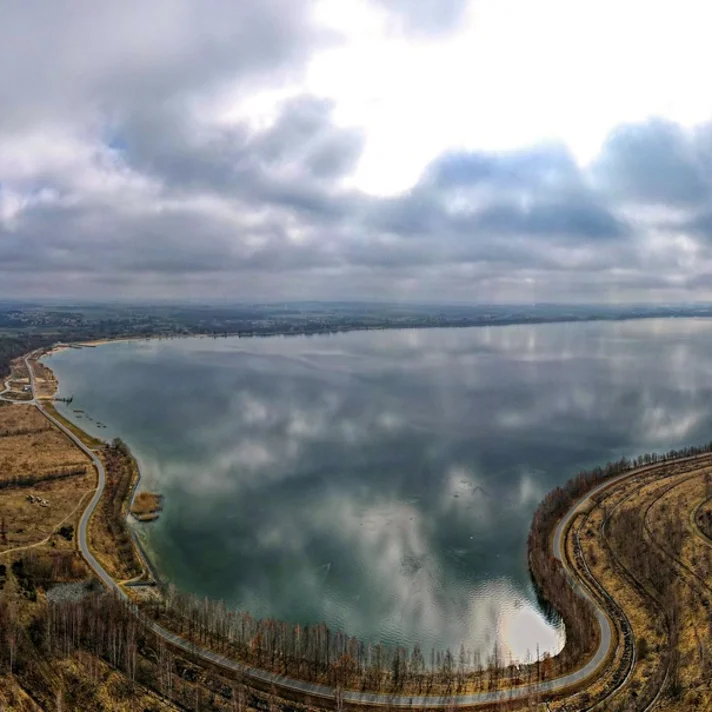 Ein foto aus der Vogelperspektive aufgenommen von einem großen See im Herbst, bewölkter Himmel, die Sonne scheint zwischen den Wolken durch.