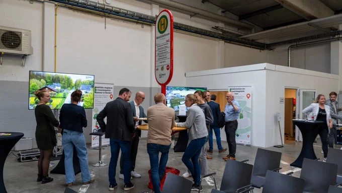 Teilnehmende unterhalten sich an einem Messestand mit Monitoren und Mobilitätsstation-Schild.