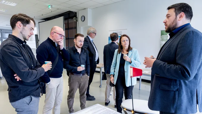 Mehrere Personen stehen in einem hellen Raum um einen Tisch mit Informationsmaterial, eine Person hält eine Kaffeetasse.